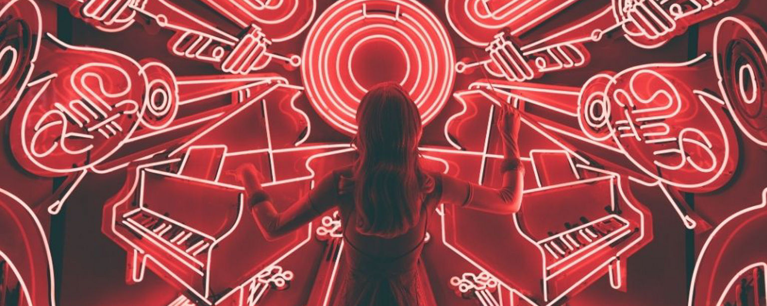 Eine junge Frau steht dirigierend vor einer Reihe rot illuminierter Musikinstrumente in Neon-Optik 